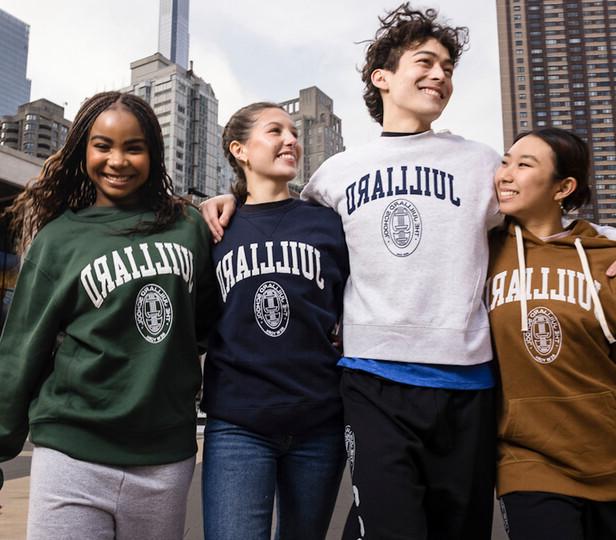 Four smiling students wearing Juilliard sweatshirts in different colors, standing arm-in-arm outdoors